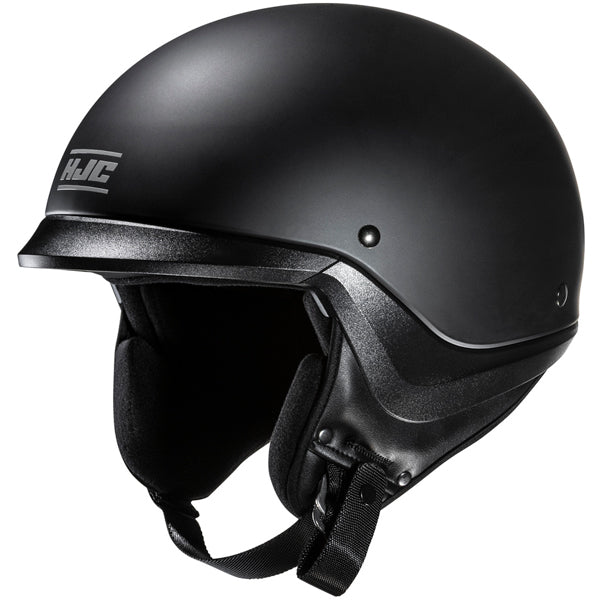 A sleek, matte black motorcycle helmet with a smooth, rounded shell and a slightly extended visor, featuring the HJC logo in white on the front left side. The interior padding is visible around the open face, made of soft black foam, and the chin strap with a quick-release buckle hangs loosely. The background is plain white, emphasizing the helmet's modern, minimalist design with subtle highlights and shadows that accentuate its contours. The image is shot from a slightly angled front view, showcasing the h