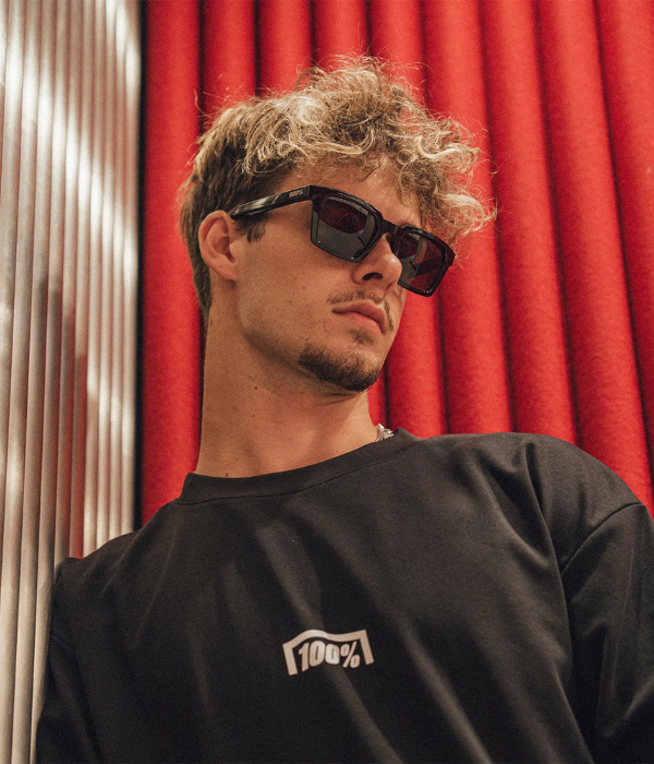 Man wearing sunglasses and a black shirt with a logo against a red curtain background 100% Goggles collection