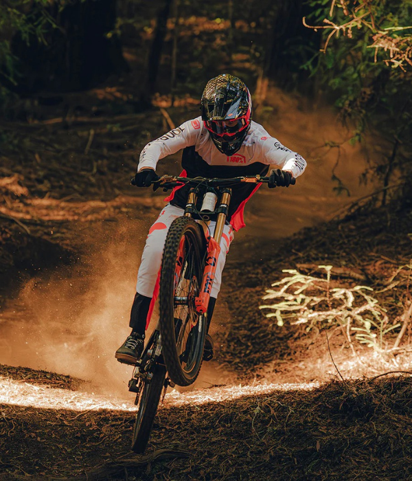 Biker in action on a dirt trail with trees and foliage in the background 100% Goggles collection