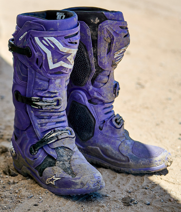 Close-up of mud-covered purple Alpinestars Tech 10 motocross boots on a dirt track after an off-road ride. collection