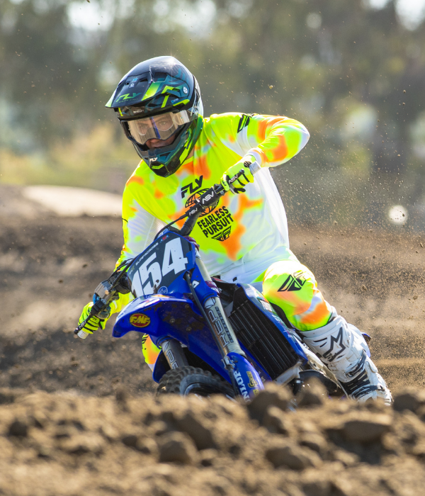 Rider wearing FLY Racing neon yellow and orange motocross gear carving a blue Yamaha dirt bike through a berm on a motocross track. collection