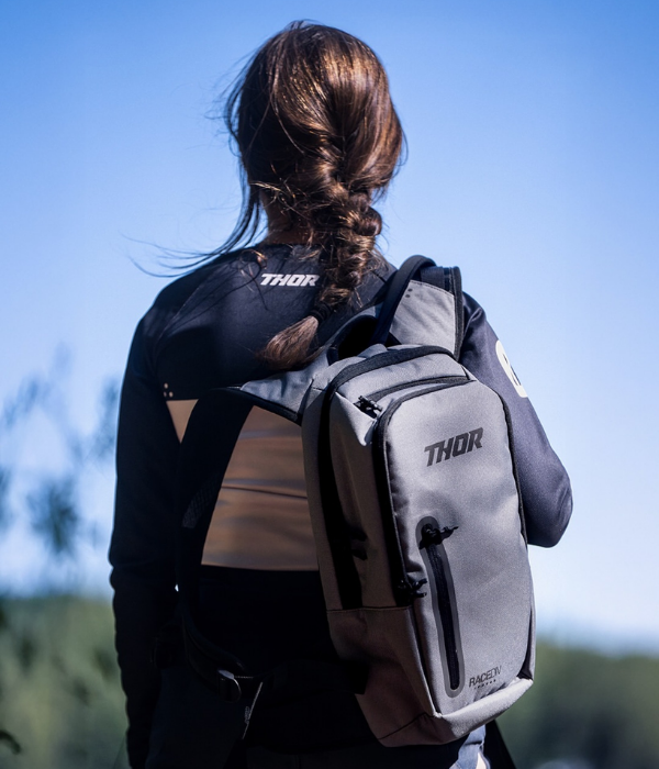 Rear view of a woman carrying a gray Thor backpack outdoors, with Thor-branded riding apparel visible against a clear blue sky. collection