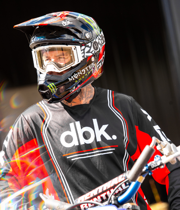 Rider wearing FLY Racing x DBK motocross gear with a custom helmet while standing beside a dirt bike in a garage setting. collection