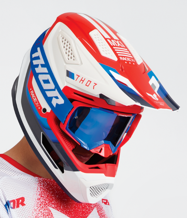 Close-up portrait of a rider wearing a red, white, and blue Thor motocross helmet with matching blue-lens goggles against a clean white background. collection