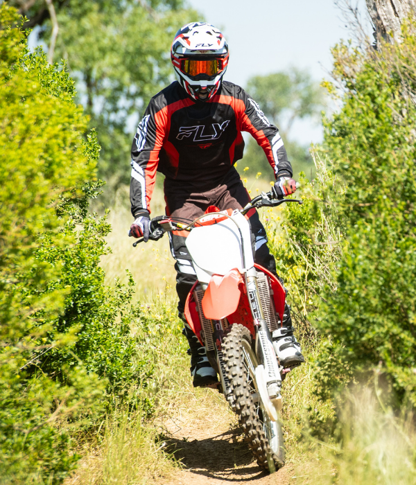 Rider wearing FLY Racing motocross gear riding a dirt bike along a narrow single-track trail through green brush. collection