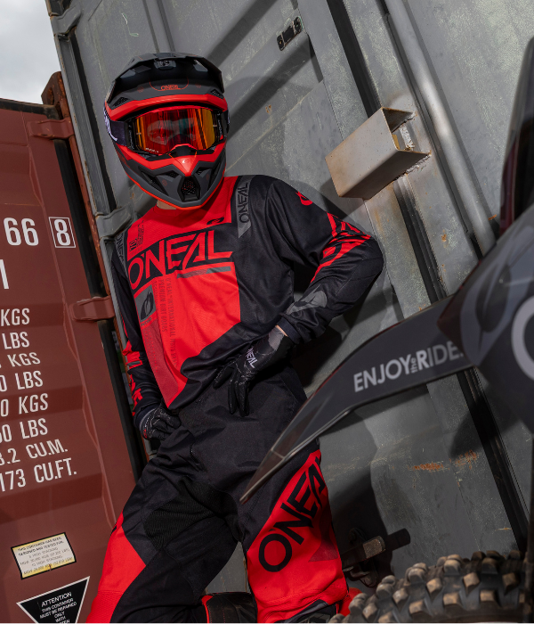 Rider wearing O'Neal black and red motocross gear standing beside a dirt bike in an industrial shipping container setting. collection