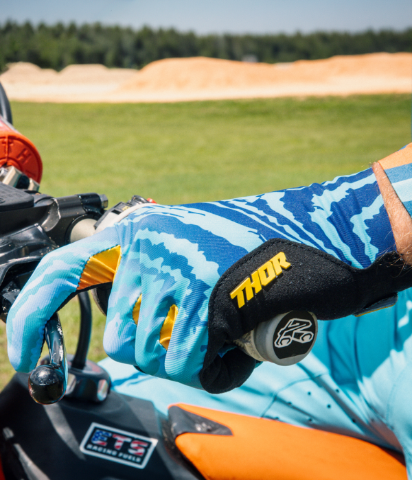 Close-up of blue patterned Thor motocross gloves gripping a dirt bike handlebar, with the throttle and controls in focus against an outdoor track backdrop. collection
