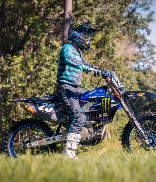 Motocross rider wearing blue and gray Thor riding gear seated on a blue Yamaha dirt bike in a grassy wooded setting. collection