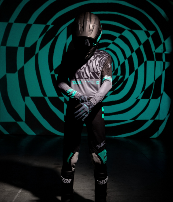 Studio portrait of a motocross rider wearing dark Thor gear with teal accents, posed against a dramatic teal-and-black geometric projection. collection