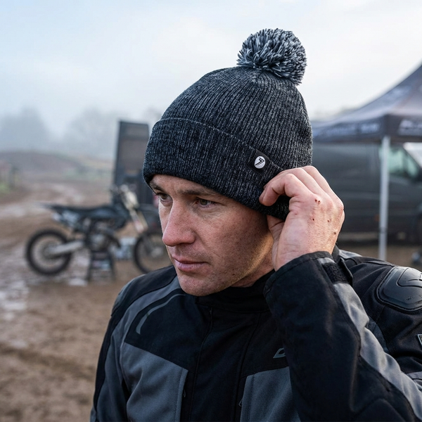 Man wearing a black beanie with a pom-pom, standing outdoors with a dirt bike and tent in the background.