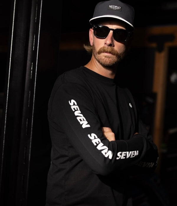 Man wearing a black cap and sunglasses, with a black long-sleeve shirt featuring 'Seven' branding. Seven MX Casual Wear collection