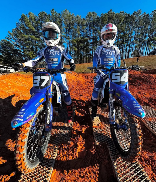 Two youth motocross riders on blue bikes lined up on a dirt track with orange clay and trees in background collection