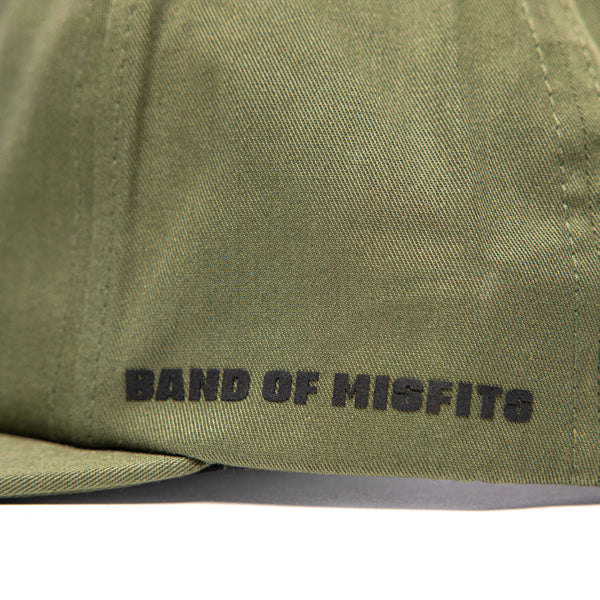 SKU 3210050 – Fasthouse Spanner Hat detail of side panel in olive green fabric with bold black “Band of Misfits” lettering, connecting motocross heritage with streetwear style.