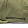 SKU 3210050 – Fasthouse Spanner Hat detail of side panel in olive green fabric with bold black “Band of Misfits” lettering, connecting motocross heritage with streetwear style.